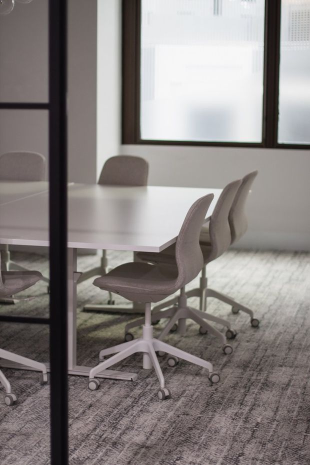 A modern meeting room viewed through a dark-framed glazed partition, featuring a large white rectangular conference table surrounded by upholstered rolling office chairs. The room has light walls, frosted privacy glazing, and textured grey carpet, creating a clean, understated commercial office setting.