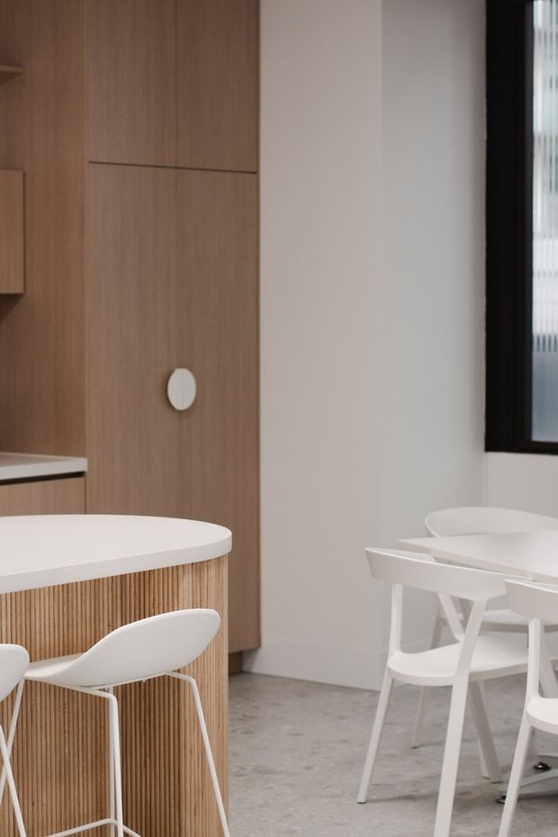 A bright, minimalist kitchen and dining area featuring a rounded white island with vertical light-wood slatted cabinetry, matching tall wood cabinetry, white bar stools, and a white dining table with chairs. The space has pale walls, a grey concrete-like floor, and a black-framed window.