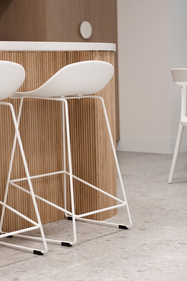 A minimalist interior detail showing two white bar stools with slim tubular frames positioned at a rounded kitchen or bar island. The island features a vertical light timber slatted front, white countertop, and warm timber cabinetry above. Pale grey tiled flooring and a neutral light grey wall complete the understated setting.