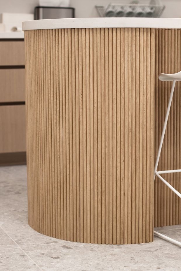 Close-up interior view of a curved kitchen island or counter clad in closely spaced vertical light timber battens, topped with a white solid-surface benchtop. Matching light timber cabinetry and pale stone-look tiled flooring are visible, with part of a white wire-frame bar stool at the right edge.