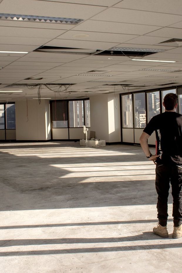 A large, vacant commercial office interior under refurbishment. The space has an exposed and damaged suspended acoustic-tile ceiling with missing panels and loose wiring, full-height internal glazed partitions and windows, a broad unfinished concrete floor, and strong sunlight casting long shadows across the room. A person stands at the right edge, likely inspecting the site. High-rise buildings are visible through the windows.
