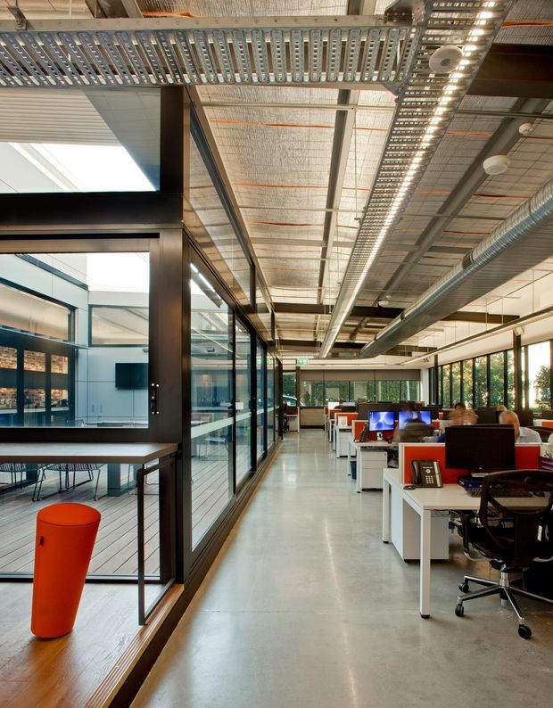 An open-plan contemporary office interior with rows of white workstations, computer monitors, office chairs and red privacy panels. A glass-partitioned meeting or breakout area runs along the left side, while large windows bring in daylight on the right. The exposed ceiling includes structural beams, cable trays, suspended acoustic panels, visible ductwork and industrial services, creating a modern industrial workplace aesthetic.