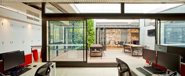 A bright contemporary open-plan office interior with workstations in the foreground opening through large glazed sliding doors onto a covered courtyard or terrace. White built-in storage lines the left wall, while timber-framed glazing, exposed brick feature walls, outdoor tables, and abundant natural light create a connection between the workspace and outdoor area. The ceiling has a linear slatted or perforated finish with integrated lighting.