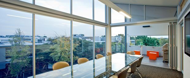A bright, contemporary open-plan dining and living space with a long glass dining table and curved tan dining chairs positioned beside floor-to-ceiling glazing. The glazing overlooks a leafy suburban setting and distant low-rise buildings. At the far end, a compact lounge or outdoor-facing area includes orange seating, while exposed ceiling beams, linear lighting, and extensive aluminium-framed glazing define the architecture.