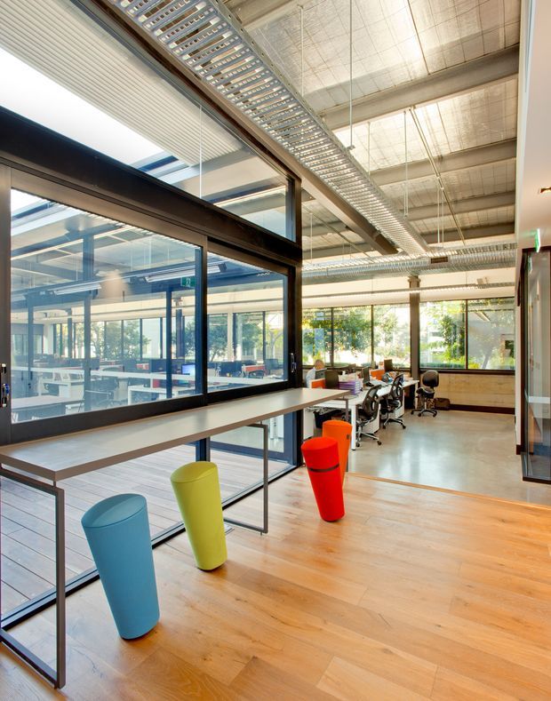 A bright, open-plan commercial office interior with a spacious circulation and breakout area. The space features exposed industrial services and ductwork, a high ceiling, extensive black-framed internal glazing, timber flooring, long built-in work counters, and colorful sculptural stools. Workstations and office furniture are visible beyond the glazed partition, with abundant natural light from large windows.