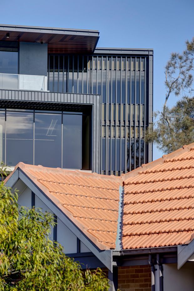 Exterior architectural photograph showing a contemporary multi-level building behind a pitched terracotta-tiled residential roof. The modern structure has dark metal framing, extensive vertical glazed openings, projecting flat roof planes, and vertical fins or battens. A glass-fronted balcony and large windows are visible, with mature vegetation at the edges and clear blue sky.