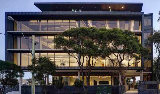 Street-facing dusk view of a contemporary multi-storey commercial or institutional building with a strongly glazed facade, dark horizontal metal framing, vertical slatted screens, and a projecting flat roof canopy. Mature sculpted trees partially screen the lower facade, with a paved pedestrian entry and low dark masonry walls at ground level.