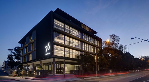 Twilight exterior photograph of a contemporary multi-storey commercial building on an urban street. The building has a dark, sharply articulated facade, extensive floor-to-ceiling glazing, illuminated interiors, projecting roof slabs, and a prominent illuminated “76” sign. Street traffic light trails and surrounding trees frame the scene.