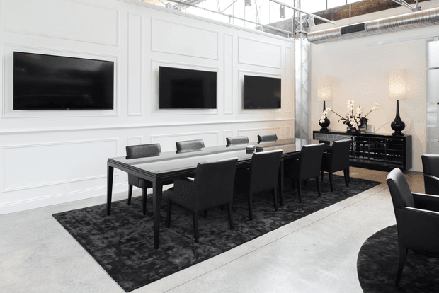 A refined open-plan dining and meeting space with a long dark rectangular table, black upholstered chairs, and three wall-mounted screens set into white decorative wall panelling. A black sideboard with lamps and floral arrangements sits against the far wall. The room combines polished classic detailing with an exposed industrial ceiling and ductwork, and has pale concrete flooring with dark patterned rugs.