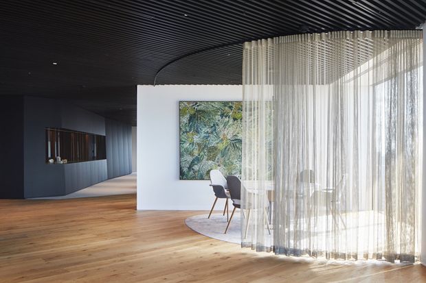 Contemporary open-plan interior with a warm timber floor, dark ribbed ceiling, and a dramatic curved sheer curtain forming a translucent dining area beside expansive glazing. A white partition wall displays artwork, while a dark built-in counter or joinery zone extends into the background. The space is refined, minimalist, and strongly contrasted, with abundant natural light.