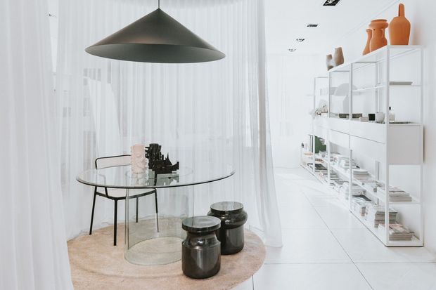 Bright minimalist dining area with a round glass dining table, slim dark metal chairs, low dark cylindrical stools, and a large charcoal pendant light. Sheer white curtains surround the space, while open white shelving displays books, ceramics, and decorative objects. The room has a white tiled floor and a soft round neutral rug.