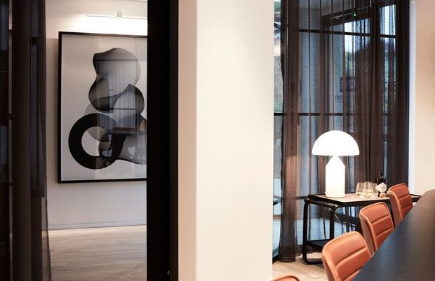 Contemporary dining and living area viewed through a wide dark-framed opening. The foreground features a long dark dining table with warm orange upholstered chairs and a white dome table lamp. Floor-to-ceiling glazing is dressed with sheer dark curtains, while an adjacent room contains a large abstract monochrome artwork and pale timber flooring.