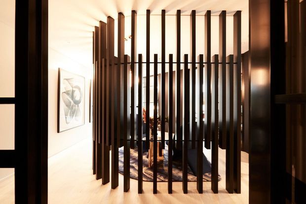Interior view of a refined contemporary space featuring a dramatic dark timber or metal vertical-slat divider. The screen partially conceals a seating area and decorative furnishings beyond, while warm concealed lighting creates strong rhythmic shadows across pale walls and flooring. Artwork is visible on the left wall, and dark framed openings or joinery define the surrounding architecture.