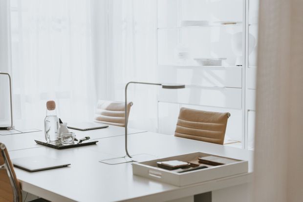 Bright minimalist white dining or work area with a large white table, tan upholstered chairs, slim linear table lamps, a serving tray, and extensive white built-in cabinetry and shelving. The space is softly lit with a clean, refined contemporary aesthetic.