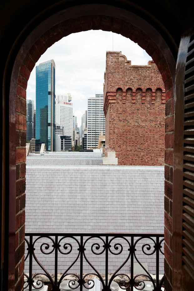View through an arched brick opening with a decorative wrought-iron balustrade, looking across a pale tiled roof toward a dense modern city skyline. The foreground architecture combines exposed red brick, a historic arched aperture, dark louvred shutters, and ornate metalwork with contemporary high-rise buildings beyond.