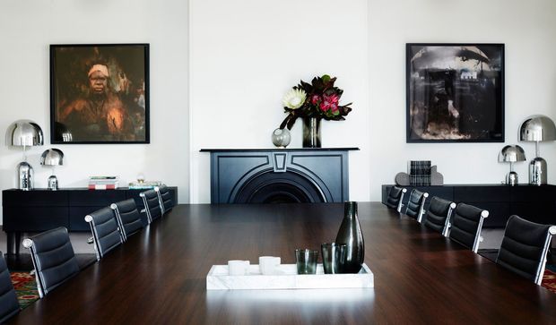 Formal dining room with a long dark timber dining table surrounded by black leather chairs, symmetrical black sideboards, chrome dome lamps, framed artwork, and a central dark decorative fireplace mantel with flowers. The room has white walls and a refined, high-contrast contemporary-classic atmosphere.