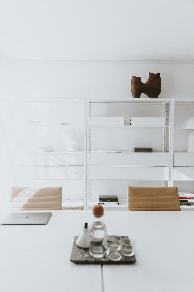 Bright, minimalist home office or dining-workspace with a large white built-in shelving and storage wall, white table, upholstered tan chairs, laptop, and a small tray of glassware. The space is predominantly white with sparse decor and strong natural light.