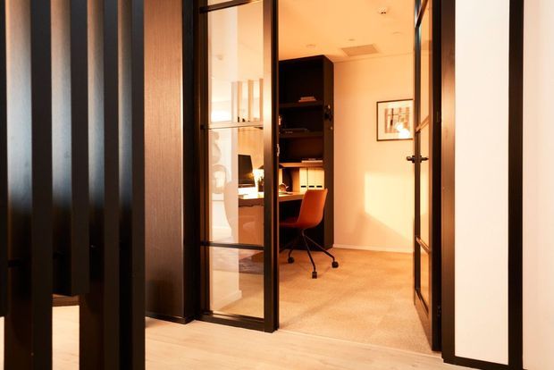 View through black-framed glazed partitions and an open glass door into a compact home office. The room contains a dark built-in desk and shelving, a burnt-orange swivel chair, filing folders, framed artwork, and warm carpet flooring, with strong afternoon sunlight creating a warm atmosphere.