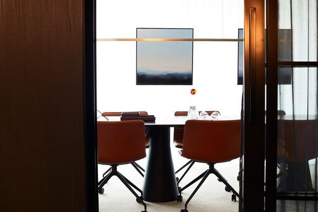 A view through a doorway into a refined dining or meeting space. A dark rectangular table is surrounded by rust-orange upholstered swivel chairs, with a black pedestal base and a light carpeted floor. Timber wall and door surfaces frame the opening, while a large bright window, horizontal timber rail, and minimalist artwork create a contemporary, warmly contrasted interior.