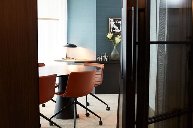 A refined dining or meeting area viewed through dark-framed glass and timber partitions. The room features a white oval table, rust-orange upholstered swivel chairs, a black desk lamp, a deep blue-green textured wall, dark timber joinery, framed artwork, and a vase of flowers. The composition has a sophisticated, moody office or dining-room character.