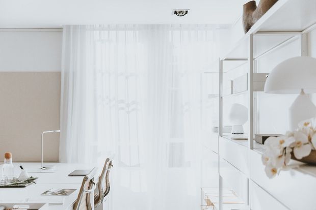 Bright, refined dining and workspace area with a long white table, slim chairs, sheer floor-to-ceiling curtains, and extensive white built-in shelving and cabinetry. Neutral beige and white finishes create an airy minimalist interior, with sculptural pendant lighting and subtle orchid decor.