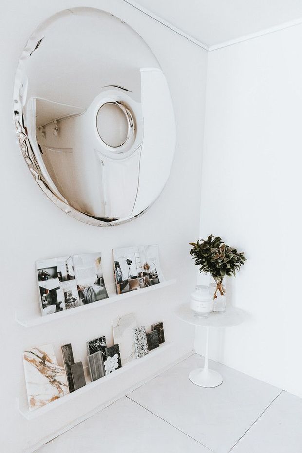 Bright, predominantly white interior corner featuring a large round wall mirror, slim picture ledges displaying books and prints, and a small white pedestal side table with a leafy plant. The mirror reflects additional white cabinetry or architectural detailing. The space has clean lines, pale tiled flooring, and a minimal gallery-like presentation.