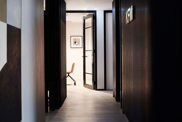 A refined interior hallway with dark timber wall panelling and vertical slatted screening, contrasting with pale walls and light timber flooring. Black-framed glazed doors open toward a small office or workspace with a peach-toned chair, while a framed artwork and illuminated wall lettering add subtle detail. The space has a contemporary, moody, high-contrast feel.