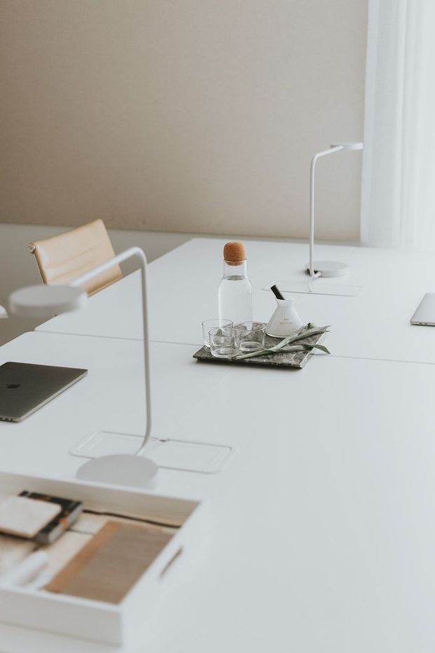 A minimalist, softly lit home office or studio workspace with a large white desk, slim white task lamps, a tan leather chair, a bottle, glassware, stationery, and open material samples. The room has a warm beige wall and bright white window treatment, creating a calm, restrained setting.