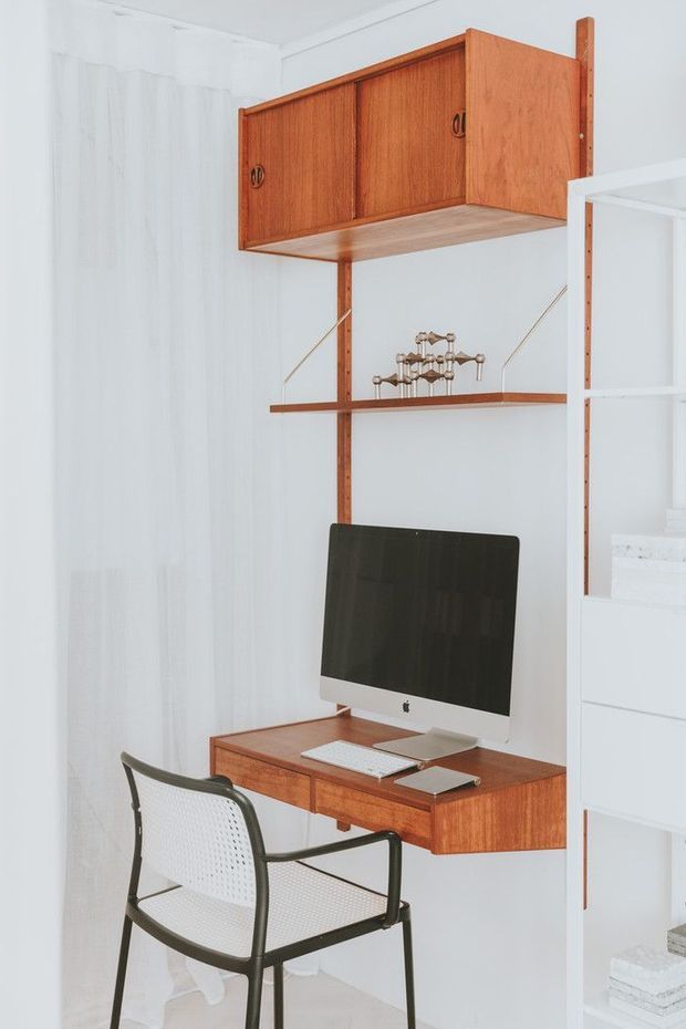 A compact built-in home workspace with a wall-mounted timber desk, matching overhead cupboards and open shelving. A desktop computer, keyboard and notebook sit on the desk, with a black-framed mesh chair in front. The cabinetry and shelving are warm reddish mid-tone timber against white walls and a white built-in shelving unit, creating a clean, space-efficient study nook.