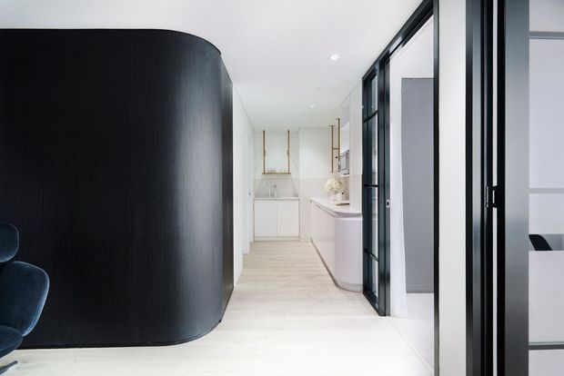 A bright, refined contemporary interior corridor leading toward a minimalist bathroom or vanity area. A dramatic curved black timber feature wall or built-in enclosure contrasts with pale timber flooring, white walls and cabinetry, and black-framed glazed doors. A partial blue upholstered chair is visible at the left edge, while the right side opens toward another light-filled space.