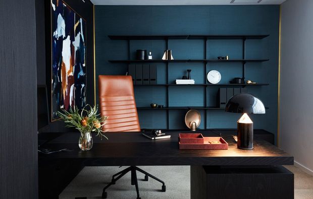A dark, refined home office with a substantial black desk, leather task chair, built-in open shelving, decorative objects, artwork, and a sculptural table lamp. The deep blue-black wall and joinery create a moody, sophisticated workspace.
