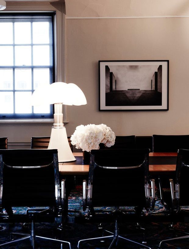 A sophisticated dining room with a long timber dining table surrounded by black leather or glossy black modern chairs. A large sculptural white table lamp and white flower arrangement sit on the table, while a framed monochrome artwork hangs on the warm beige wall. A tall gridded window with dark timber framing provides natural light, and a patterned rug is visible beneath the table.