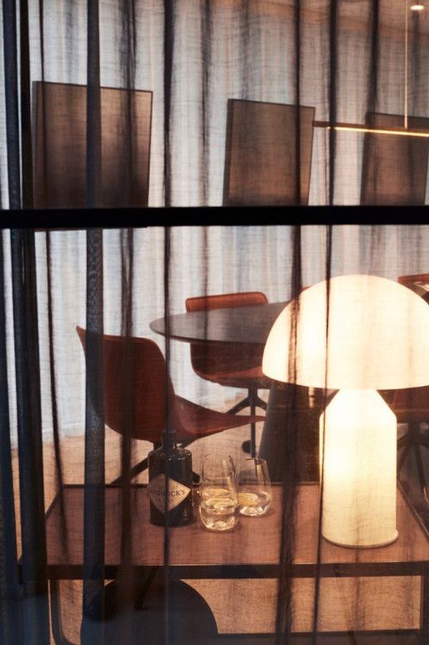Moody interior view through sheer black curtains, showing a warm timber dining area with a dark oval table, sculptural wooden chairs, and a glowing table lamp on a console. Black-framed glazing or partitions create strong horizontal and vertical lines, while translucent curtains add texture and privacy.