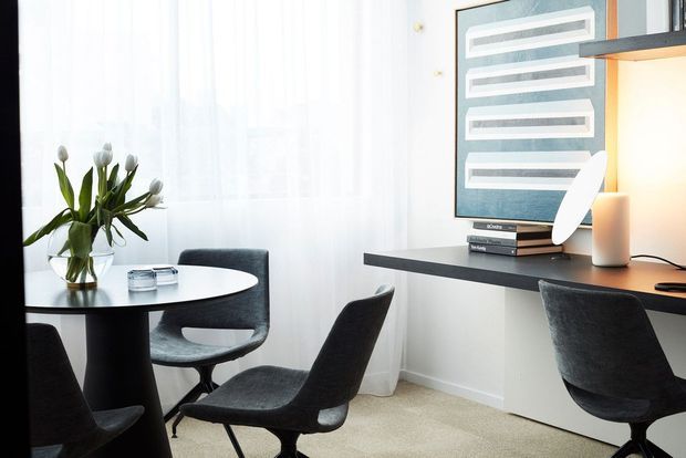 Bright, refined home office and dining nook with a dark built-in desk, computer monitor, upholstered office chair, round dining table, black chairs, sheer curtains, and a vase of tulips. A framed abstract artwork and warm desk lighting add contrast to the light neutral interior.