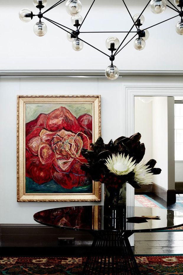 A refined interior vignette featuring a round black glossy console table with a sculptural base, a tall dark floral arrangement, and a large ornate-framed floral painting. The room has pale grey-white paneled walls, dark timber flooring, patterned area rugs, and a striking contemporary black linear chandelier with spherical glass globes. A bright adjoining room is visible through a wide trimmed opening.