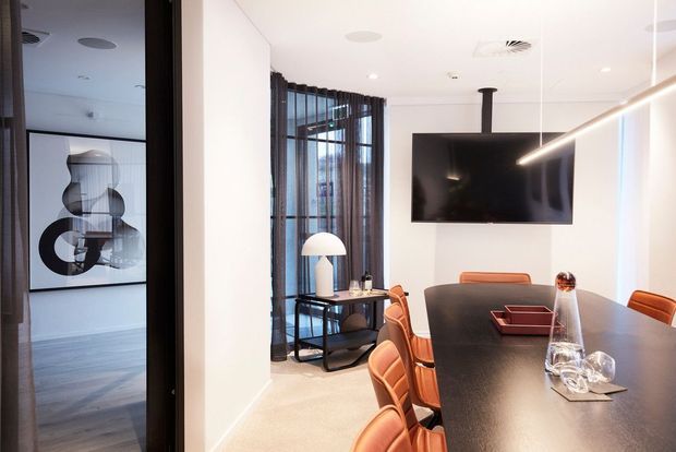 Contemporary open-plan dining and living area with a large dark dining table, rust-toned upholstered dining chairs, a wall-mounted television, linear pendant lighting, and a compact black console beneath full-height glazed doors with sheer curtains. A hallway or adjacent room with abstract artwork is visible through the opening on the left.