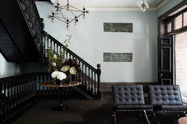A refined, moody entry hall with a dramatic black-painted staircase and balustrade, pale blue-grey walls, dark carpet, and a black-framed glazed entry door. Two black tufted leather lounge chairs sit in the foreground beneath sculptural globe chandeliers, while a small round console table displays flowers. Decorative wall plaques and traditional trims add character.