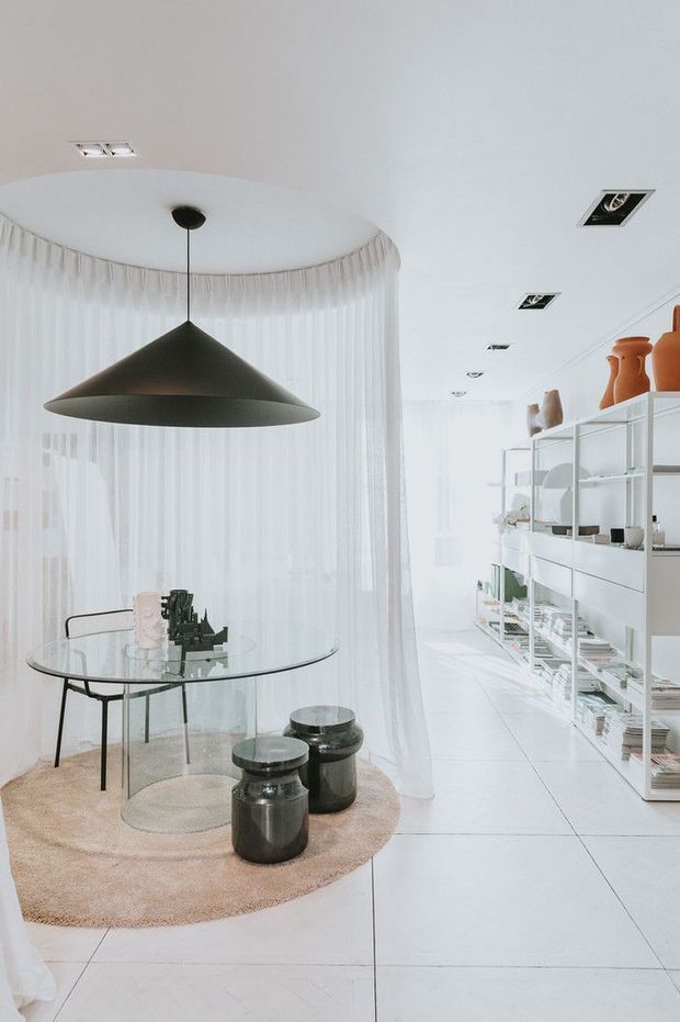Bright, refined open-plan dining and display area with sheer floor-to-ceiling curtains, a sculptural dark pendant light, round glass dining table, black chairs, and dark cylindrical stools on a pale round rug. White tiled flooring, white walls, and open built-in shelving filled with ceramics and books create a minimalist gallery-like interior.