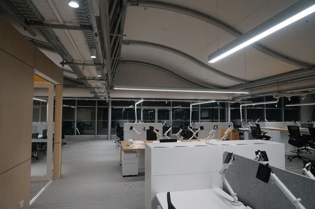 Open-plan commercial office interior with rows of workstations, white desks, black office chairs, glass partitions, exposed ceiling services, and linear LED lighting. The space has a clean, contemporary workplace fit-out with a neutral grey and white palette.