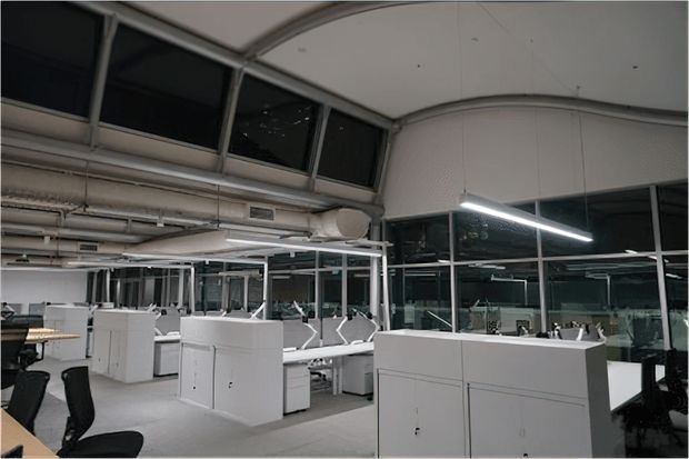 An open-plan commercial office interior with rows of white workstation partitions and desks, black office chairs, extensive internal glazing, exposed services and ductwork, and linear ceiling lighting. The space has a functional, contemporary workplace fit-out with a neutral grey-and-white palette.