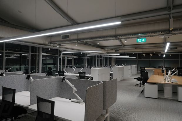 An open-plan commercial office with rows of workstations, grey acoustic privacy screens, white desks, black office chairs, and a larger timber meeting or collaboration table. The space has exposed services, suspended linear LED lighting, extensive internal glazing or reflective partitions, and a neutral grey-and-white fit-out.