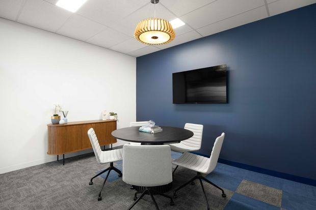 A contemporary office meeting room with a round dark table, four white upholstered office chairs, a wall-mounted television or presentation screen, and a timber credenza with small plants. The room has a deep navy accent wall, white walls, grey carpet with blue and grey patterned sections, a suspended ribbed pendant light, and a suspended grid ceiling with recessed lighting.