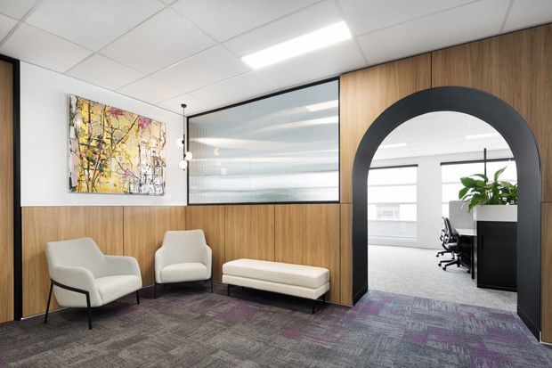 A contemporary commercial office reception or waiting area with warm wood wall panelling, grey patterned carpet with purple accents, white lounge chairs and ottoman, and a large black arched opening leading to an open-plan office. Glazed partitions, recessed linear lighting, and abstract wall art complete the space.