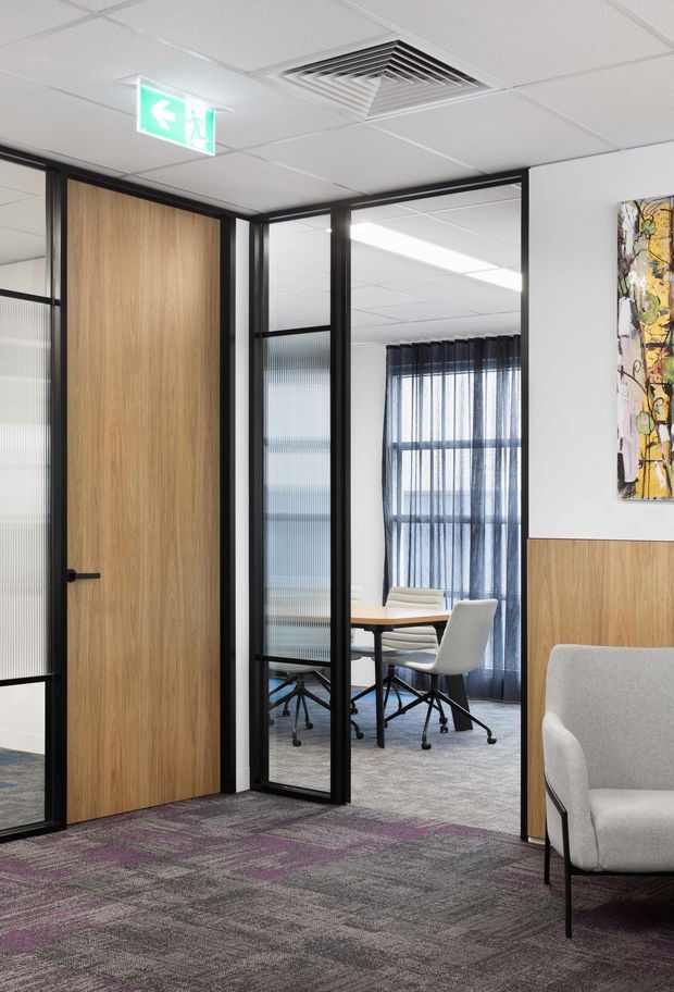 Modern commercial office interior viewed from a reception or circulation area, with black-framed glazed partitions, a timber veneer door and wall panel, carpet tile flooring, a meeting room with a round table and office chairs, acoustic curtains, and a contemporary lounge chair. Suspended acoustic ceiling tiles, linear lighting, and an illuminated exit sign reinforce the workplace setting.