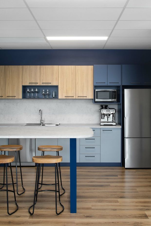 A compact contemporary kitchen with a light timber and muted blue cabinetry scheme. The kitchen features a pale stone island benchtop with blue support legs, integrated open glassware shelving, stainless-steel appliances, a concrete-look splashback, timber flooring, and a suspended acoustic ceiling with recessed fluorescent lighting. Wooden bar stools provide seating at the island.