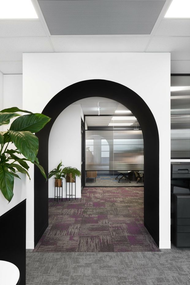 A contemporary commercial office interior viewed through a large black-trimmed arched opening. The foreground has grey carpet, white walls, indoor plants, and a partial black reception or storage unit. Beyond the arch is a glazed meeting or workspace with patterned purple-grey carpet, desks, office chairs, and suspended ceiling lighting. The space has a clean, high-contrast, modern office fit-out.