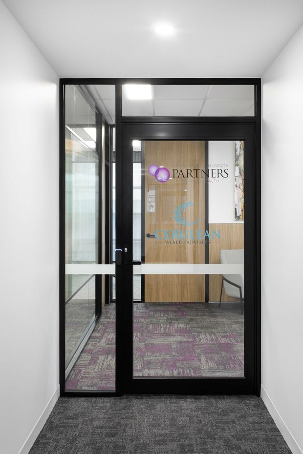 A narrow commercial office entry hall leading to a black-framed glazed door and sidelights. White walls and ceiling contrast with dark patterned carpet flooring, while the glazing reveals a reception or waiting area with timber-look wall panelling, branded signage, and a chair beyond.