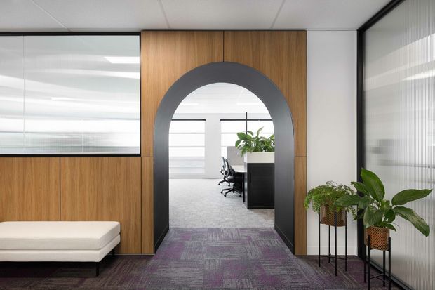 Contemporary office interior featuring a warm timber-panelled partition with a large dark arched opening, glazed office walls, carpeted circulation space, a built-in upholstered bench, and indoor plants. A workstation area is visible through the archway.