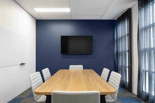A contemporary meeting room with a large light-wood conference table surrounded by upholstered white chairs. The room has a deep navy feature wall with a wall-mounted screen, whiteboard panels, a suspended white ceiling with recessed lighting, and full-height windows covered by dark sheer curtains.