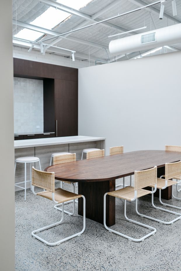 Bright open-plan commercial interior with a large dark timber dining or meeting table surrounded by woven cane chairs with white tubular metal frames. A compact kitchenette with a light stone or concrete-look island and dark timber cabinetry sits behind the table. The space has a high exposed white-painted industrial ceiling with skylights, visible structural beams, suspended services, and a large cylindrical HVAC duct. The floor is light grey terrazzo, and the overall palette is white, grey, dark timber, and natural cane.